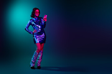 Obraz premium Full body portrait of stunning young woman hold phone empty space wear shiny dress neon light dark blue background