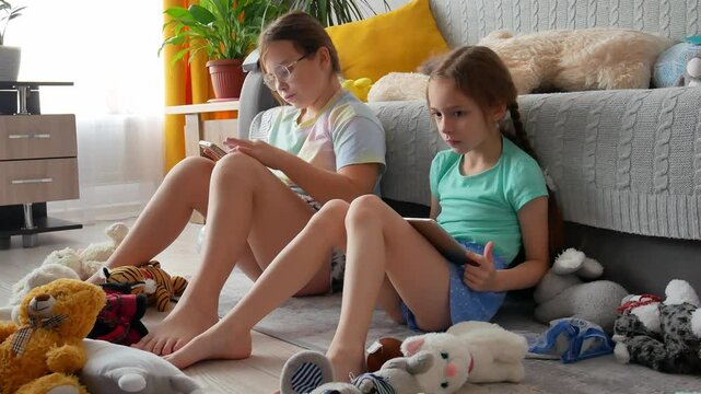 Two sisters are sitting on the floor among soft toys, holding gadgets, a phone, a tablet and playing video games. A little girl rubs her nose, has a runny nose. On the eldest daughter.