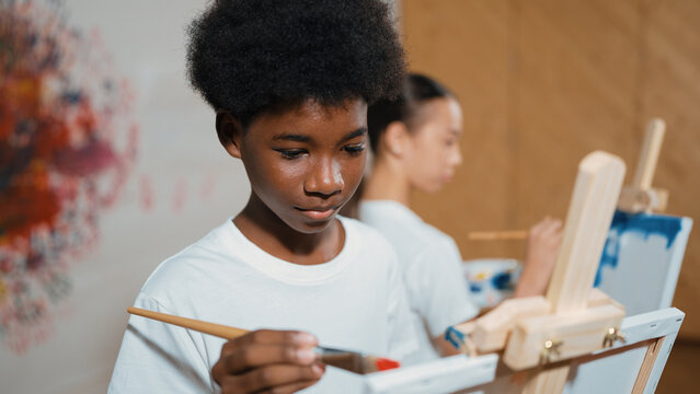 African boy painting canvas with watercolor while happy caucasian girl draw cool tone picture at colorful stained wall. Multicultural highschool student attend creative activity together. Edification.