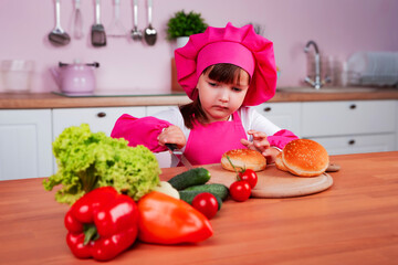 Serious little beautiful girl in a chef costume prepares burgers while sitting at a table in the kitchen. Healthy eating.