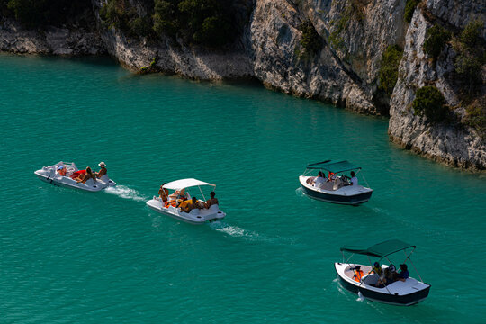 Tourists in four little electric pleasure boats and pedalos or pedal boats on turquoise water of Verdon Gorge, exploring the valley and river on hot summer day, with majestic cliffs.
