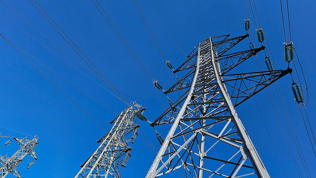 Low angle view looking up at metal electricity pylons against a clear bright blue sky; electricity pylons or towers with cables and wires; concept electricity transmission, electrical infrastructure.