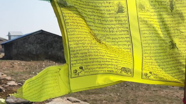 A yellow buddhist and tibet prayer flag is waving in the foreground. In the background there is a traditional house on a trekking path on the Himalayan mountain range