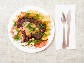Dinner with premium t bone steak, fried potatoes chips with ridge, fresh salad with feta cheese on white plate and light color table cloth. Large portion of food with high quality products.