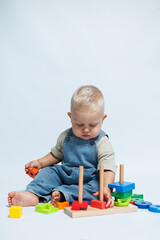 Cute blond boy in dungarees sits on a white background and plays with a wooden sorter. Sorter for the development of fine motor skills of babies. Child plays with educational toys.
