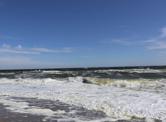 Fototapeta premium Foamy waves of the Black Sea under a clear blue sky. A serene view of the Black Sea shoreline with foamy waves crashing onto the sandy beach, framed by a bright blue sky and gentle clouds
