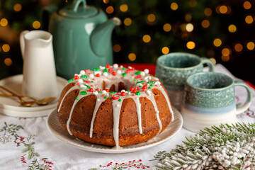 Festive table with bundt cake with Christmas sprinkles and powdered sugar icing. Tea pot and mugs. New Year tree on background.