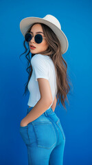 Fototapeta premium Full-length portrait of a beautiful fashion model posing on a bright blue background. Dressed in a white tank top, jeans, and a gray hat, she stylishly holds round sunglasses 