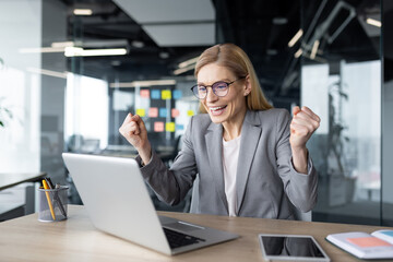 Fototapeta premium Mature businesswoman expressing excitement as she celebrates a work success. The image captures her joy and enthusiasm in a modern office setting while working on her laptop.