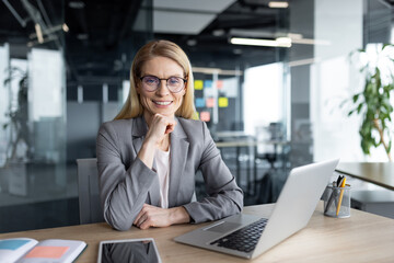 Fototapeta premium Mature woman businesswoman smiling confidently while working on a laptop in a modern office. Her professional demeanor and stylish attire reflect success and leadership qualities.