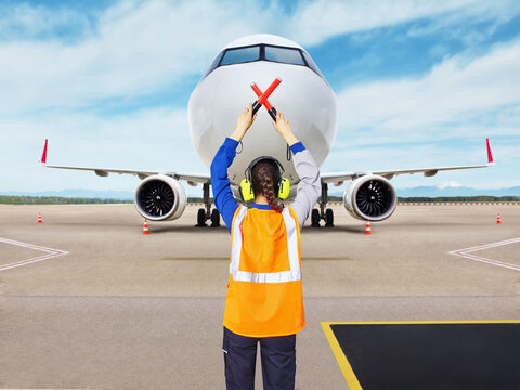 Airport ground staff member wearing a safety vest and headphones signals an approaching airplane with wands on the runway during aircraft marshalling procedure. Concept of aviation safety