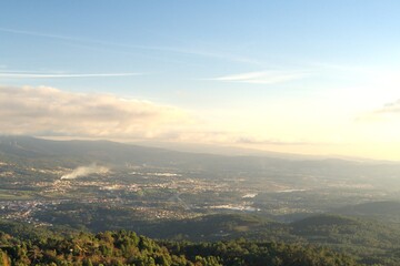 The image shows a panoramic view of a town in a valley surrounded by hills and mountains. The foreground has a dense forest, and buildings and roads are visible, suggesting early morning light.