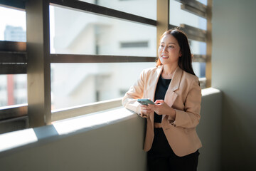 Young asian businesswoman wearing beige jacket is holding smartphone and looking out window in office building, enjoying sunlight and urban view