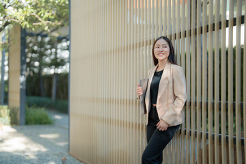 Young asian businesswoman is holding a laptop and leaning against a wooden wall outdoors, enjoying the fresh air and natural light while working remotely