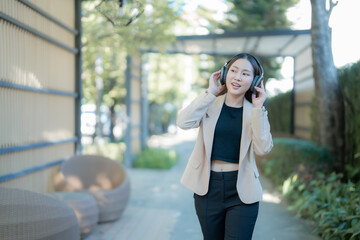 Fototapeta premium Young asian businesswoman wearing wireless headphones enjoying music while walking outdoors in a modern urban environment, combining work-life balance with technology and leisure