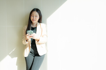Asian businesswoman is using smartphone and leaning against wall in modern office building with natural sunlight coming through window, creating bright and productive atmosphere