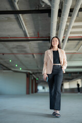 Obraz premium Smiling businesswoman walking through a parking garage, holding a smartphone while heading to work or returning home after a busy day, exuding confidence and professionalism