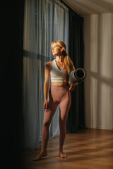 A blonde woman holding a yoga mat stands in a spacious studio by the window, bathed in the rays of the setting sun.