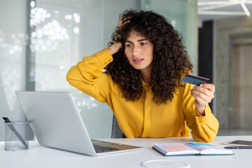 Obraz premium A worried businesswoman holds a credit card while using her laptop, indicating online financial issues or shopping problems. Her expression suggests stress or surprise, reflecting financial concerns.
