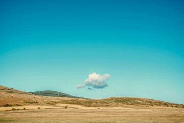 A single white cloud floats in a clear blue sky above a peaceful, golden countryside landscape. Minimalist and tranquil scene.