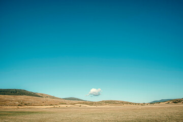 A single white cloud floats in a clear blue sky above a peaceful, golden countryside landscape. Minimalist and tranquil scene.