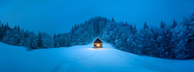 Panorama of picturesque winter landscape with glowing wooden cabin in snowy forest. Cozy house in...