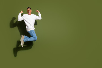 Young man in white sweater and jeans jumps joyfully against a khaki background, expressing happiness and excitement in casual fashion style