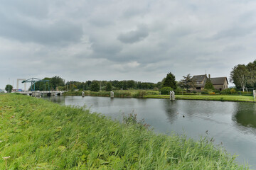 Green grass along a tranquil canal with a bridge and house under overcast skies in a peaceful rural...