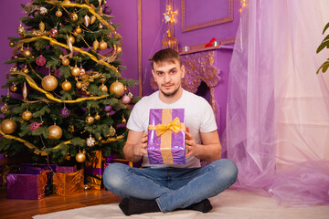 young man in white t-shirt and jeans on purple christmas background holding gift box . christmas tree with toys