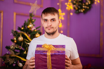 young man in white t-shirt and jeans on purple christmas background holding gift box . christmas tree with toys