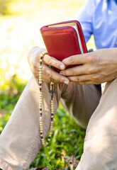 An unrecognizable man holds a Bible and a Catholic rosary while standing outdoors, symbolizing union with God and freedom during Holy Week