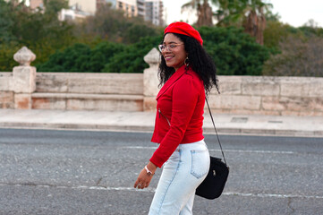 Stylish young woman with curly hair, wearing a red jacket, beret, and jeans, walks confidently along a city street, carrying a black bag and smiling