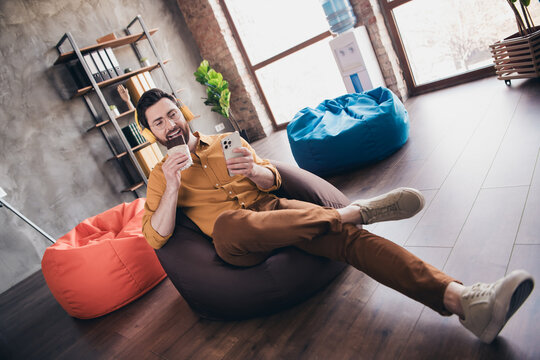 Full body photo of man sit beanbag chocolate gadget listen music wear yellow shirt formalwear modern office room interior indoors workspace