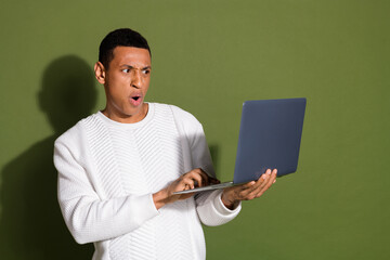 Young stylish man holding laptop with surprised expression against khaki background in casual white...