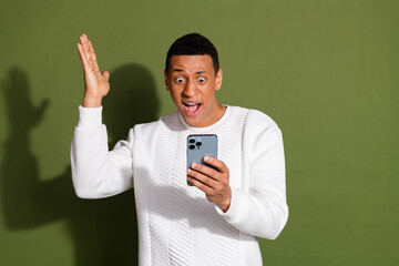 Excited young man in white sweater using smartphone against a khaki background, expressing surprise...