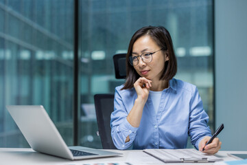 Asian woman in a modern office, concentrating on her laptop taking notes. This image captures...