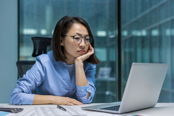 Focused Asian businesswoman seated at a desk, concentrating on laptop screen in a office...