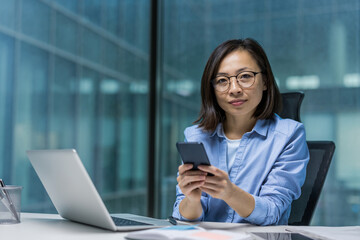 An Asian businesswoman sits at her desk, using a smartphone with confidence. She embodies professionalism and focus in a modern office setting, emphasizing work and technology.