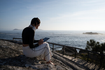 Young woman with bare feet relaxes with her tablet, watching the sunset on a rock by the ocean, creating a peaceful image of work life balance