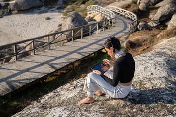 Fototapeta premium Young woman in casual attire sits barefoot on a coastal rock, using a tablet with a wooden footbridge in the background