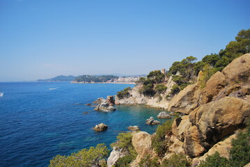 Fototapeta premium Mediterranean coastline with rugged cliffs, turquoise water, pine trees, and a distant view of a coastal town under a clear blue sky.