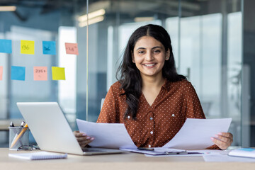 A confident businesswoman sitting at a desk, holding documents and smiling at the camera. She is...