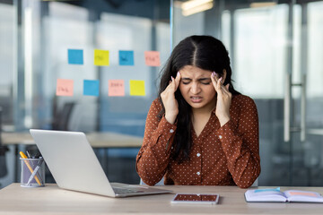 A stressed woman in an office setting, feeling a headache while working on a laptop with a...