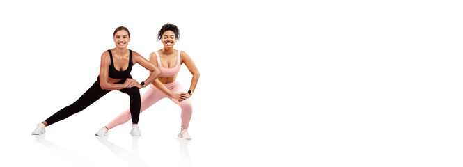Two women perform side lunges together in a bright, minimalistic indoor space. They wear matching athletic attire and demonstrate camaraderie while focusing on their fitness routine.
