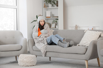 Frozen young woman in winter clothes with plaid lying on sofa at home
