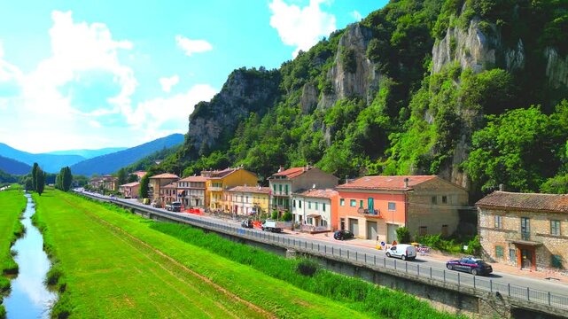 Sublime aerial pedestal up shot from Pioraco with the light-mirroring, reflective waters of the Potenza River enclosed between green shores, a nearby road with diverse vehicles, Sibillini mountains