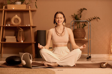 Beautiful young woman meditating with laptop at home in evening
