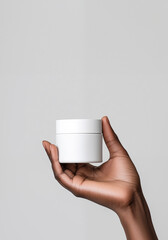 Product photography of a hand holding a small white jar, against a plain background, with an African American skin. White cosmetic lotion mockup. Flat lay; front view; copy