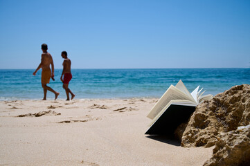 An open book lies against a stone on a sandy shore. Two men walk along the shore. Behind them is the vast ocean.