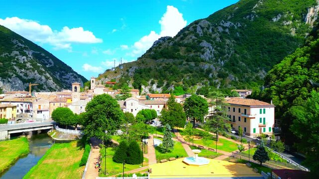 Special aerial truck left shot from Pioraco, featuring the park, trees, Potenza river, bridge, road and cars, buildings and belltowers, the backdrop of impressive Sibillini mountains, puffy cloudy sky